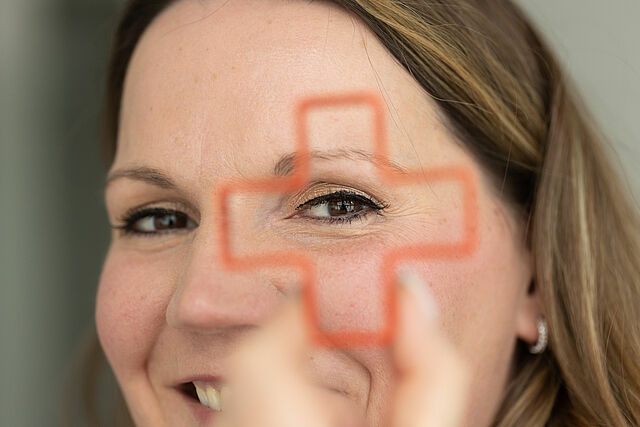 Frau hält sich ein rotes Kreuz aus Draht vor ein Auge und schaut lächelnd hindurch